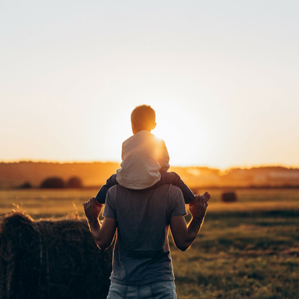 A man is holding his child on his shoulders as they watch the sun go down in a field.