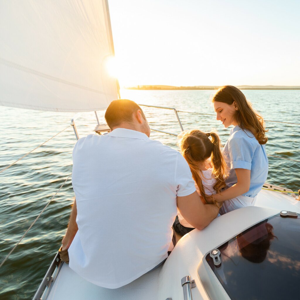 A mother and father are sitting with their daughter near the hatch of a boat. The sun is shinning down on them.