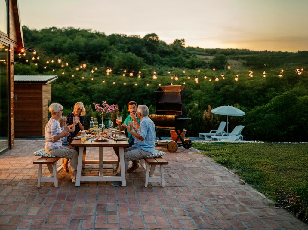 A family are sitting in the garden enjoying an evening meal. There is a barbecue in the background and rolling hills.
