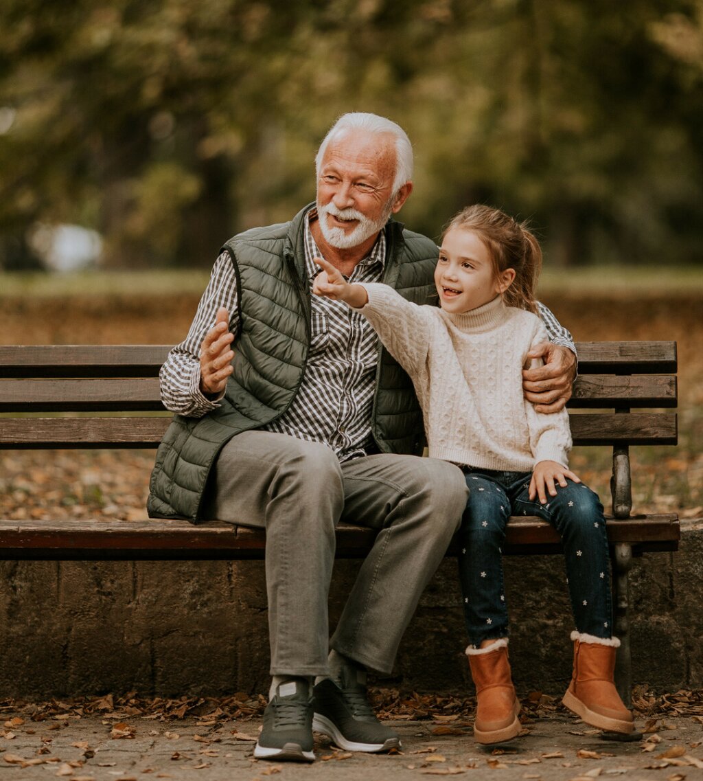 A grandfather and his grandaughter are sitting on a park bench in autumn.