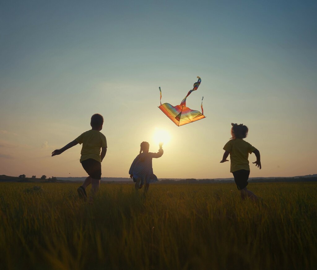 3 children are running thorugh a field in the sunset while flying their kite.