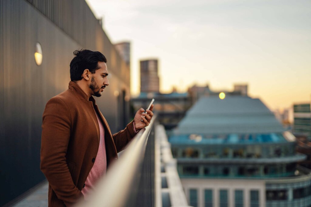 Man on a balcony at sunset, looking at his phone. The city skyline is in the background.